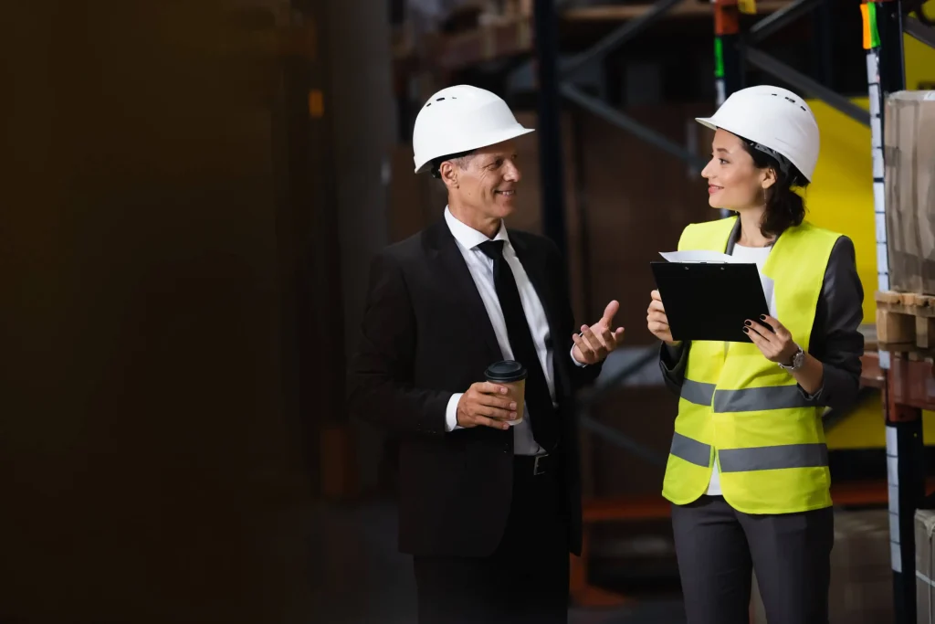 In a bustling warehouse, a business executive and a safety manager engage in a productive discussion, emphasizing the importance of safety protocols and project updates while dressed in professional and safety gear.